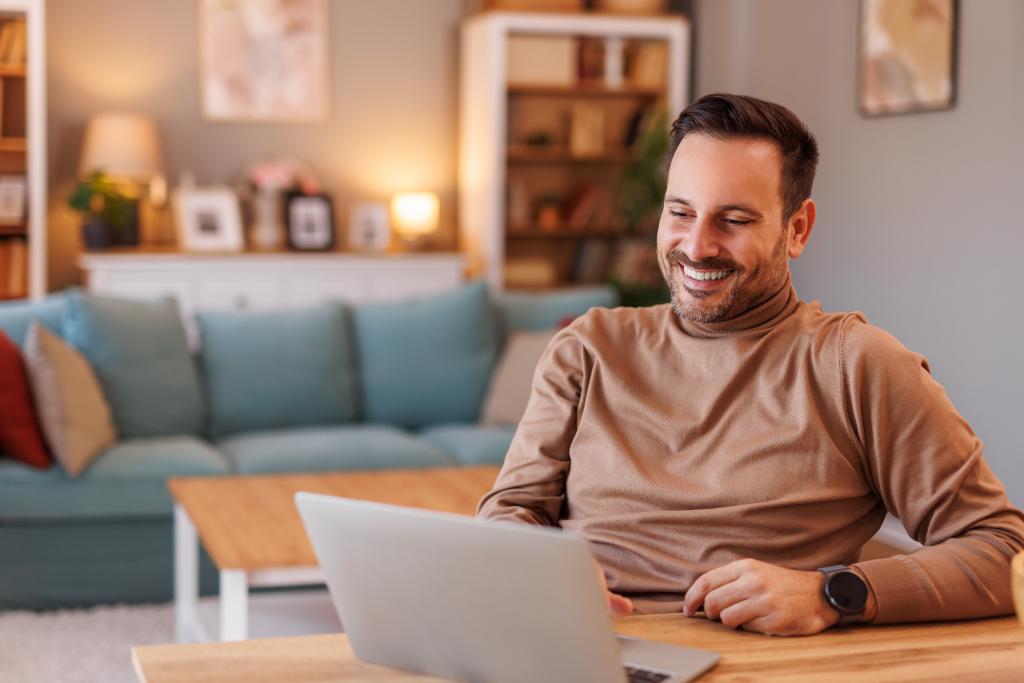 Mensch sitzt am Tisch mit Laptop vor einem gemütlichen Wohnzimmer im Hintergrund