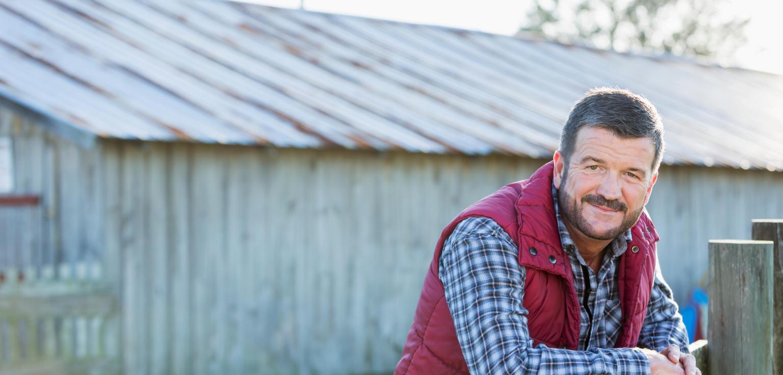 Mensch sitzt draußen an einem Zaun mit einer alten Scheune im Hintergrund