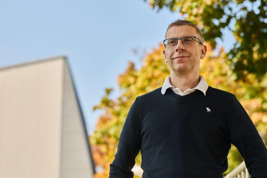 Michael Steiner steht im Freien vor einem modernen Gebäude mit bunten Herbstbäumen im Hintergrund.