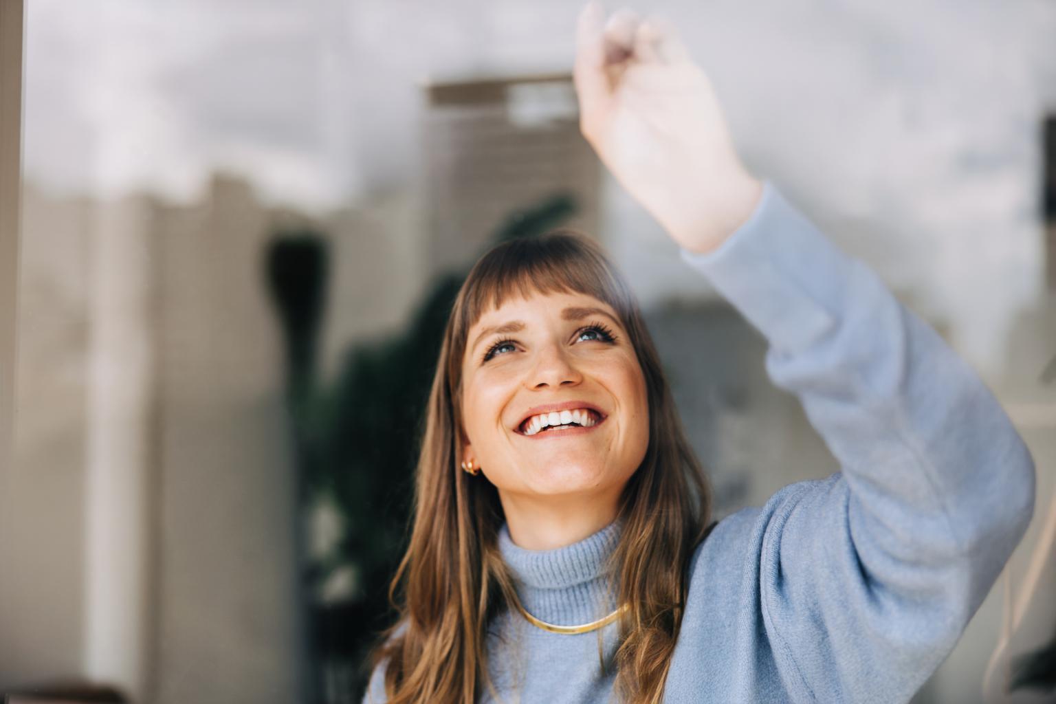 Eine Person winkt im Büro vor einem Fenster, hinter ihr reflektiert sich die Umgebung.