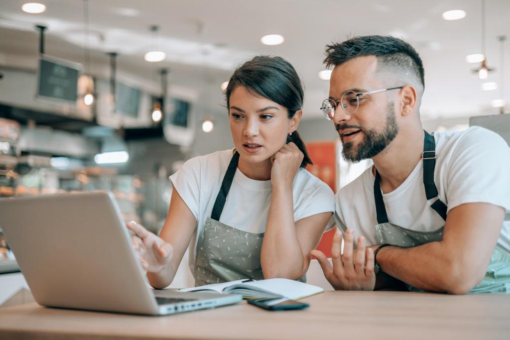 Zwei Personen in Schürzen, die über einen Laptop in einem Café-Setting diskutieren.