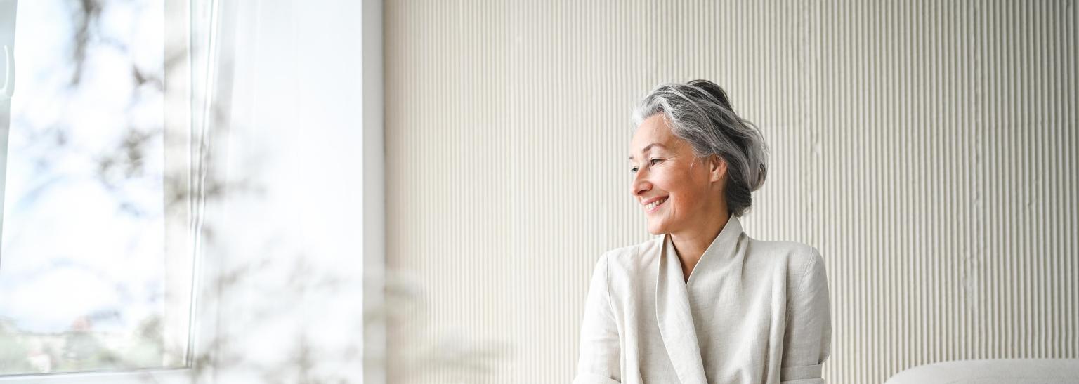 Ältere Frau sitzt entspannt im hellen Raum vor einem Fenster mit Sicht nach draußen