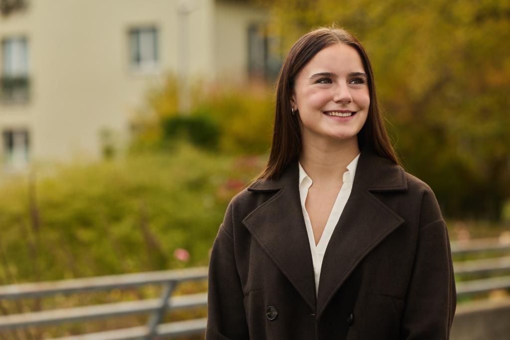 Sienna Gschwind in dunklem Mantel vor Naturhintergrund bei Tageslicht.