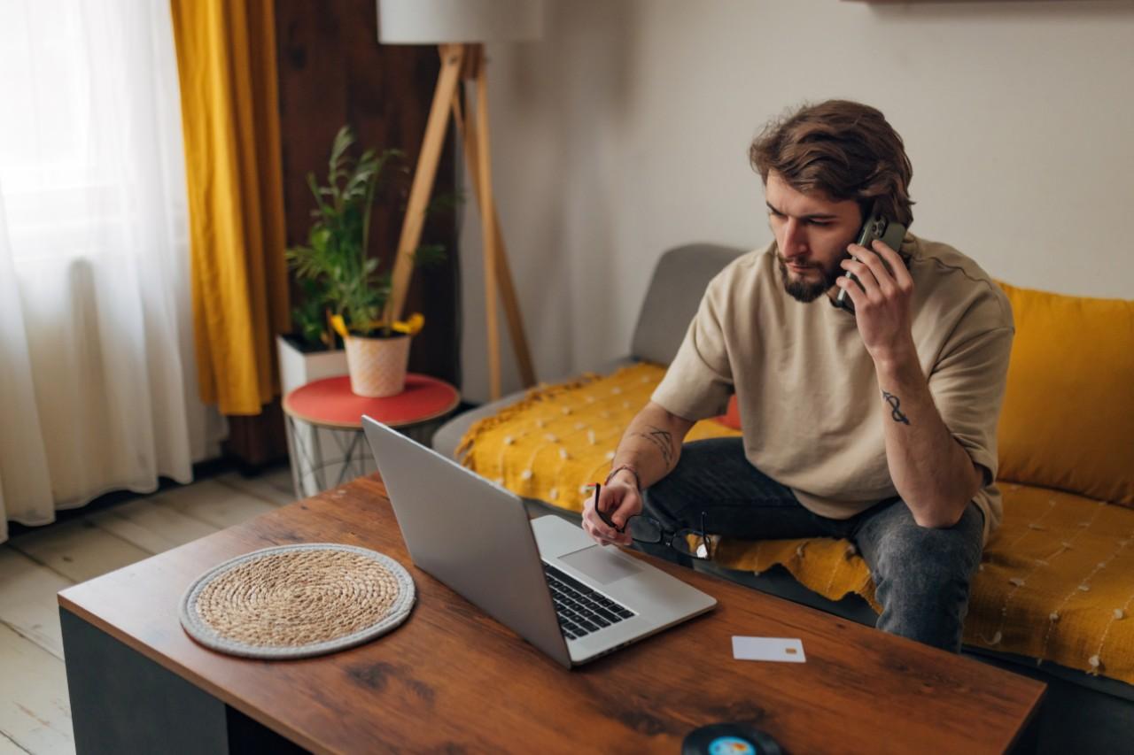 Ein Mann sitzt an einem Tisch und benutzt einen Laptop, während er am Telefon spricht, in einem gemütlichen Wohnzimmer. aus dem Artikel Wie kann ich mich effektiv vor Online-Betrügern schützen?