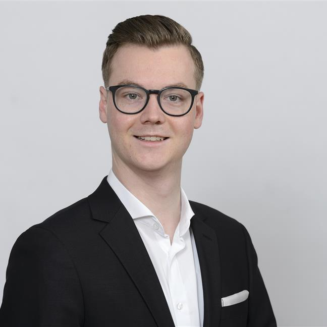 Ein junger Mann in einem Anzug mit Brille posiert selbstbewusst vor einem neutralen Hintergrund.
