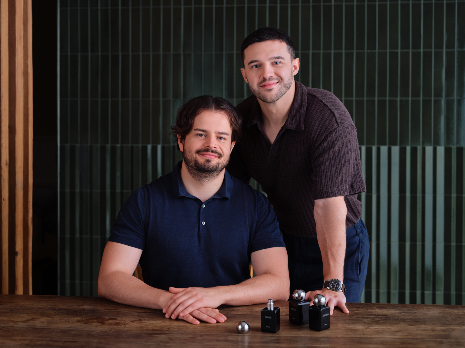 Zwei Männer sitzen an einem Holztisch vor einem grünen Gitter.