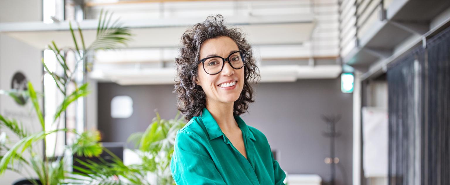 Frau steht in modernen Büro mit Pflanzen, BLKB Logo im Hintergrund.