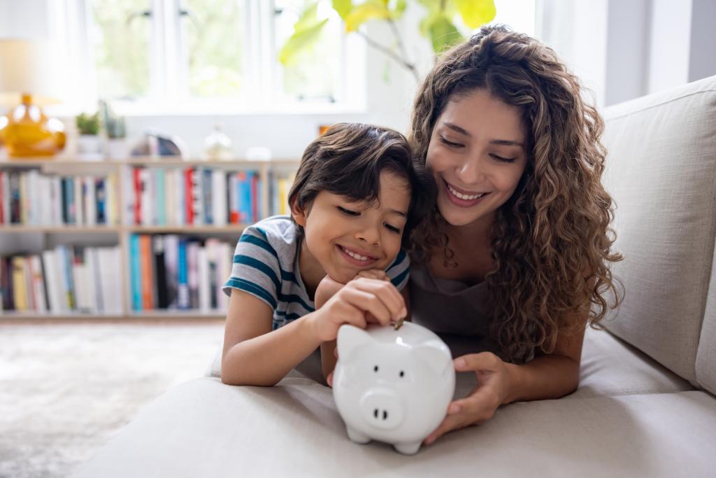 Ein Kind und eine Frau setzen Münzen in eine Sparfigur in einem Wohnzimmer