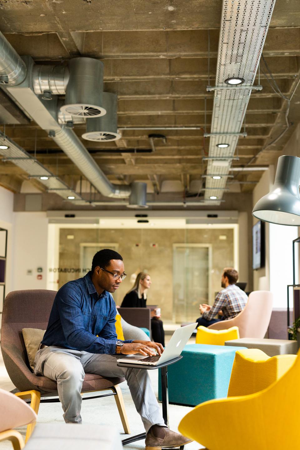 Person sits on armchair with a desk pulled up working on a laptop, two people chat in the background