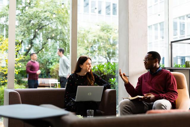 Two people sat down, one holds a laptop and the other holds a notebook, there are two people outside in the background talking