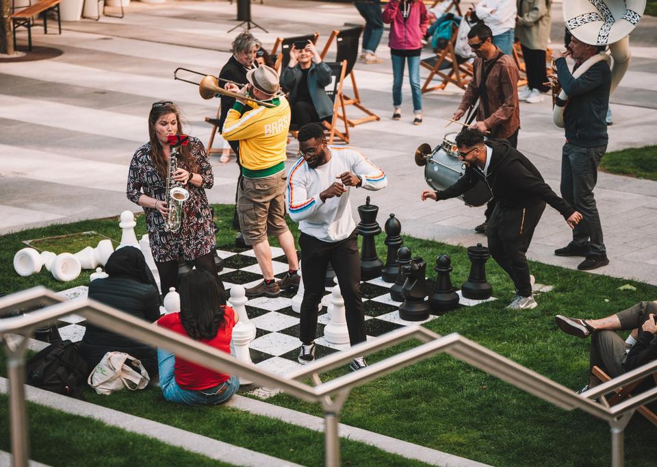 A group of musicians performing to an audience while people dance on a giant chess set.