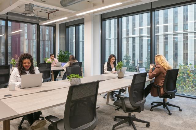 People work at their desks on laptops which are decorated with green plants, in an office with floor to ceiling windows overlooking Symphony Park