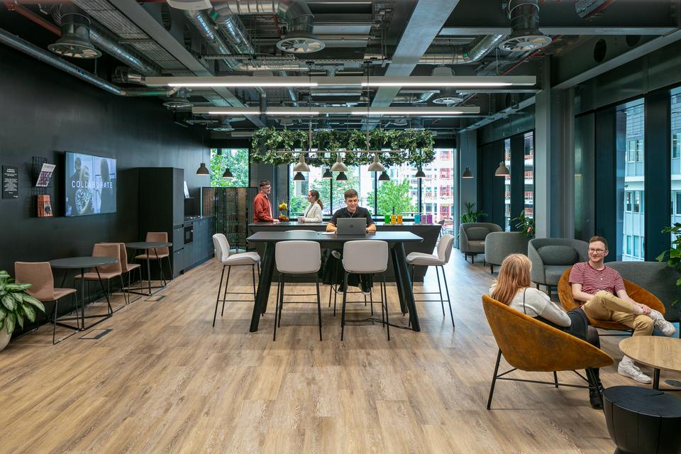 Person working on their laptop at a high table while two sit in egg chairs talking, and two people use the shared kitchen