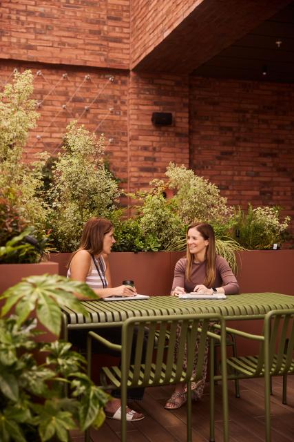 Man and a woman sat at a desk outside smiling while working on a laptop