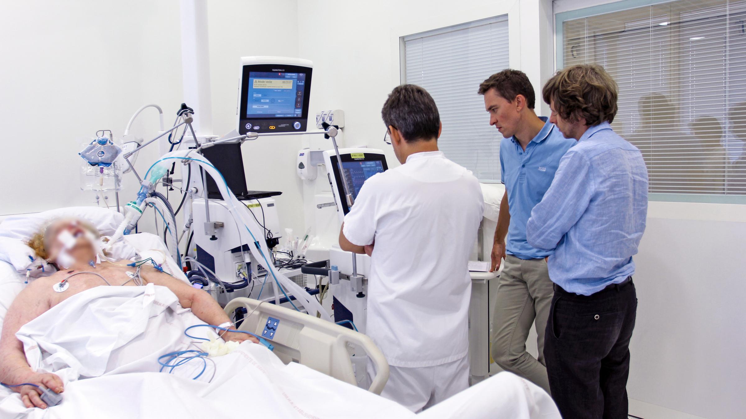 Intubated patient lies on the left side of the bed. Several people are standing next to a ventilator on the right side.