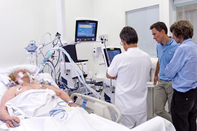Intubated patient lies on the left side of the bed. Several people are standing next to a ventilator on the right side.