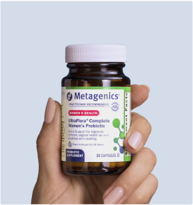 Hand holding a bottle of Metagenics UltraFlora® Complete Probiotic in front of a light blue background. The label calls out 9-in-1 daily support for digestion, skin health, immune health, and metabolism, with a clear view of the amber capsules inside.