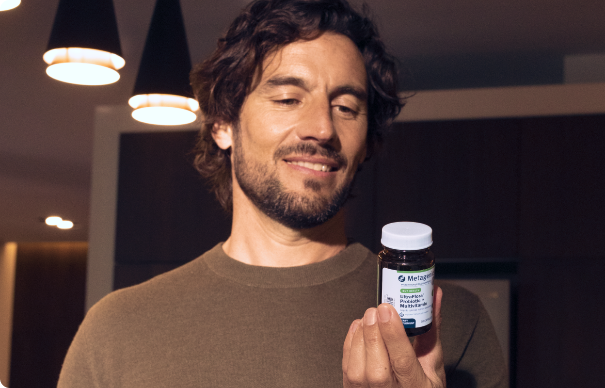Smiling man with a beard holding a Metagenics UltraFlora Probiotic + Multivitamin bottle in a warmly lit modern room, reading the label.