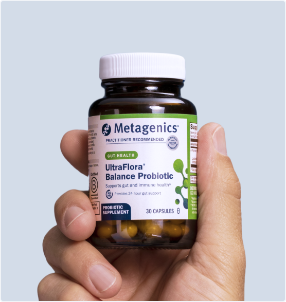 Hand holding a bottle of Metagenics UltraFlora® Balance Probiotic against a light blue background. The label highlights gut health and immune support, with a white cap and visible amber capsules inside the clear bottle.