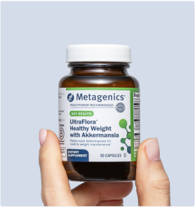 Hand holding a bottle of Metagenics UltraFlora® Complete Probiotic in front of a light blue background. The label calls out 9-in-1 daily support for digestion, skin health, immune health, and metabolism, with a clear view of the amber capsules inside.