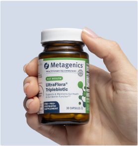 Hand holding a bottle of Metagenics UltraFlora® Complete Probiotic in front of a light blue background. The label calls out 9-in-1 daily support for digestion, skin health, immune health, and metabolism, with a clear view of the amber capsules inside.