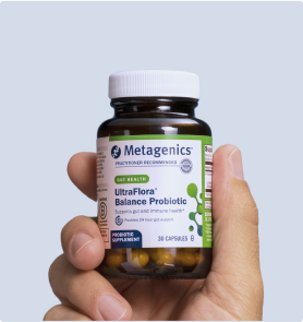 Hand holding a bottle of Metagenics UltraFlora® Complete Probiotic in front of a light blue background. The label calls out 9-in-1 daily support for digestion, skin health, immune health, and metabolism, with a clear view of the amber capsules inside.