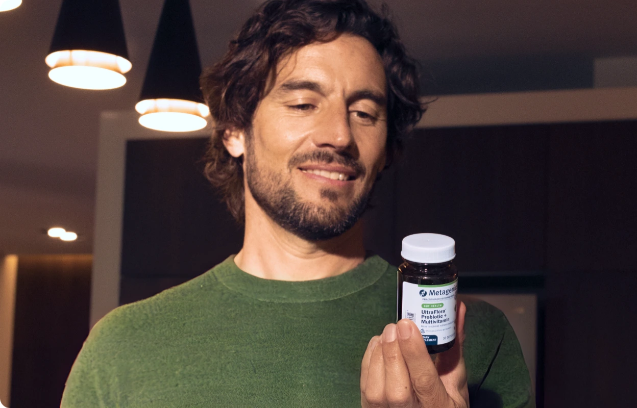 Smiling man with a beard holding a Metagenics UltraFlora Probiotic + Multivitamin bottle in a warmly lit modern room, reading the label.