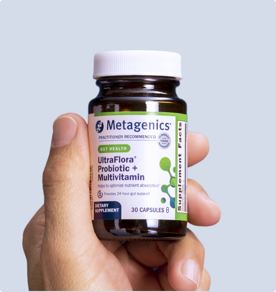 Hand holding a bottle of Metagenics UltraFlora® Probiotic + Multivitamin against a light blue background. The label emphasizes 24-hour gut support and nutrient absorption, with the Supplement Facts panel partially visible on the right.

