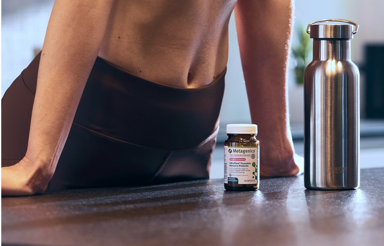 Close-up of a woman in athletic leggings with midriff exposed, leaning against a kitchen countertop beside a stainless steel water bottle and a glass bottle of Metagenics UltraFlora® Complete Women's Probiotic. The focus on her core emphasizes gut health and an active, wellness-oriented lifestyle, reinforcing the value of personalized probiotic support.