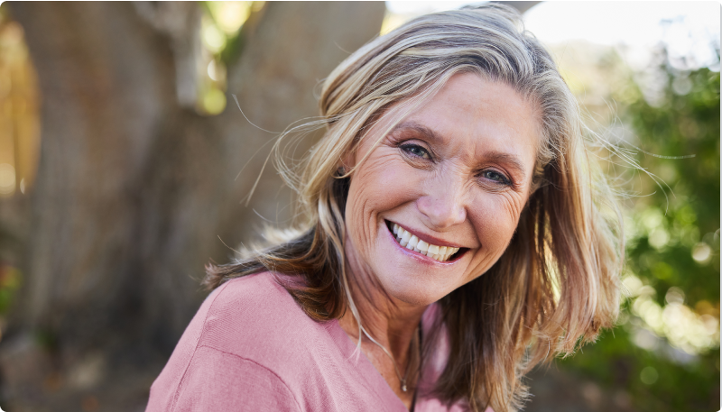 A woman with short hair wearing a pink top sits outdoors surrounded by trees and greenery. The image accompanies an article about natural solutions for hormonal balance, sleep, and stress.