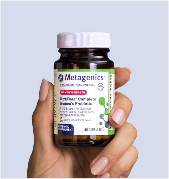 A hand with manicured nails holding a bottle of Metagenics UltraFlora® Complete Women's Probiotic against a light blue background. The label highlights 5-in-1 daily support for digestive, immune, vaginal, and emotional health, and bloating.