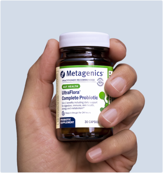 Hand holding a bottle of Metagenics UltraFlora® Complete Probiotic in front of a light blue background. The label calls out 9-in-1 daily support for digestion, skin health, immune health, and metabolism, with a clear view of the amber capsules inside.
