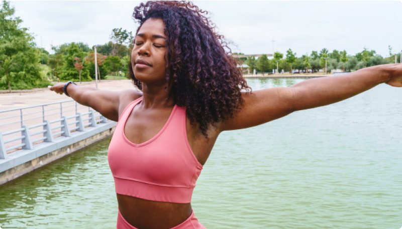 A woman wearing a pink sports bra and matching leggings stands outdoors near a body of water with her arms outstretched. The background features greenery and a paved walkway. The image accompanies an article about how gut health impacts women’s hormones and mood.