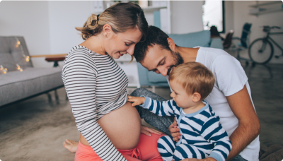A pregnant woman sits on a couch in a cozy living room, with a man kneeling beside her and a young child gently touching her belly. The background includes home furniture and a bicycle. The image accompanies an article about vital nutrients and reflects how HerWellness™ can support hormonal balance through all stages of a woman’s life, including pregnancy.