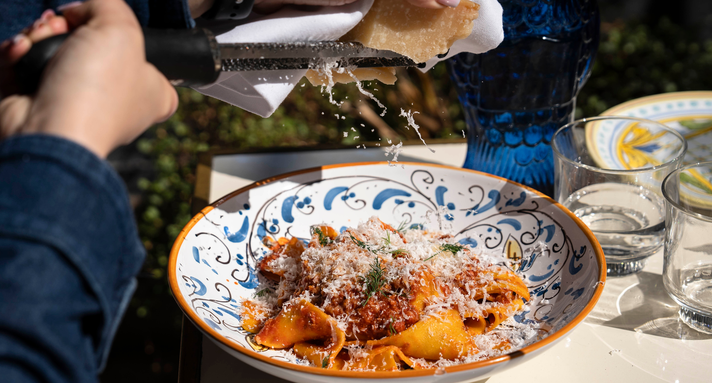 Mains râpant du parmesan au-dessus d’un bol de pâtes italiennes et motif de vaisselle rehaussant le charme du plat.