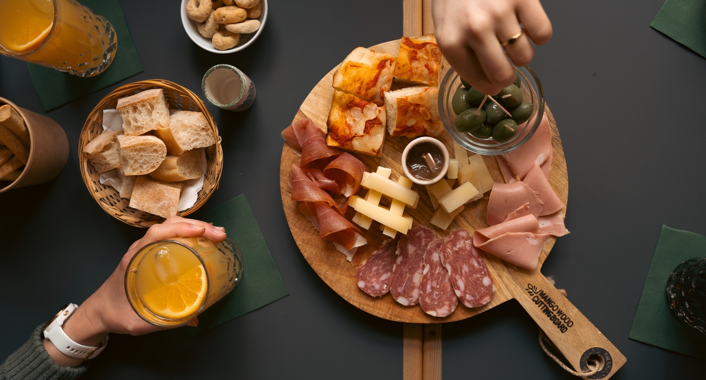Auf einem Tisch liegt eine Servierplatte mit verschiedenen Fleischsorten, Oliven, Käse und Gebäck belegt. Daneben liegt ein Körbchen mit Brot und weiterem Gebäck, sowie Getränke in Gläsern.