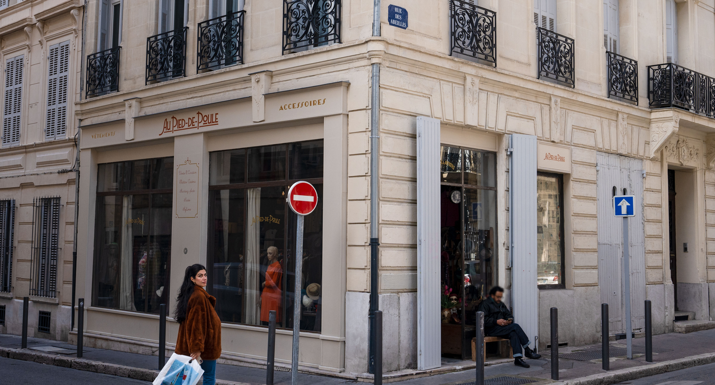 Eine Passantin geht an dem Eckladen 'Au Pied de Poule' vorbei, einem Geschäft für Secondhand-Mode in Marseille, mit einer Vielzahl von Kleidungsstücken sichtbar durch das Schaufenster.