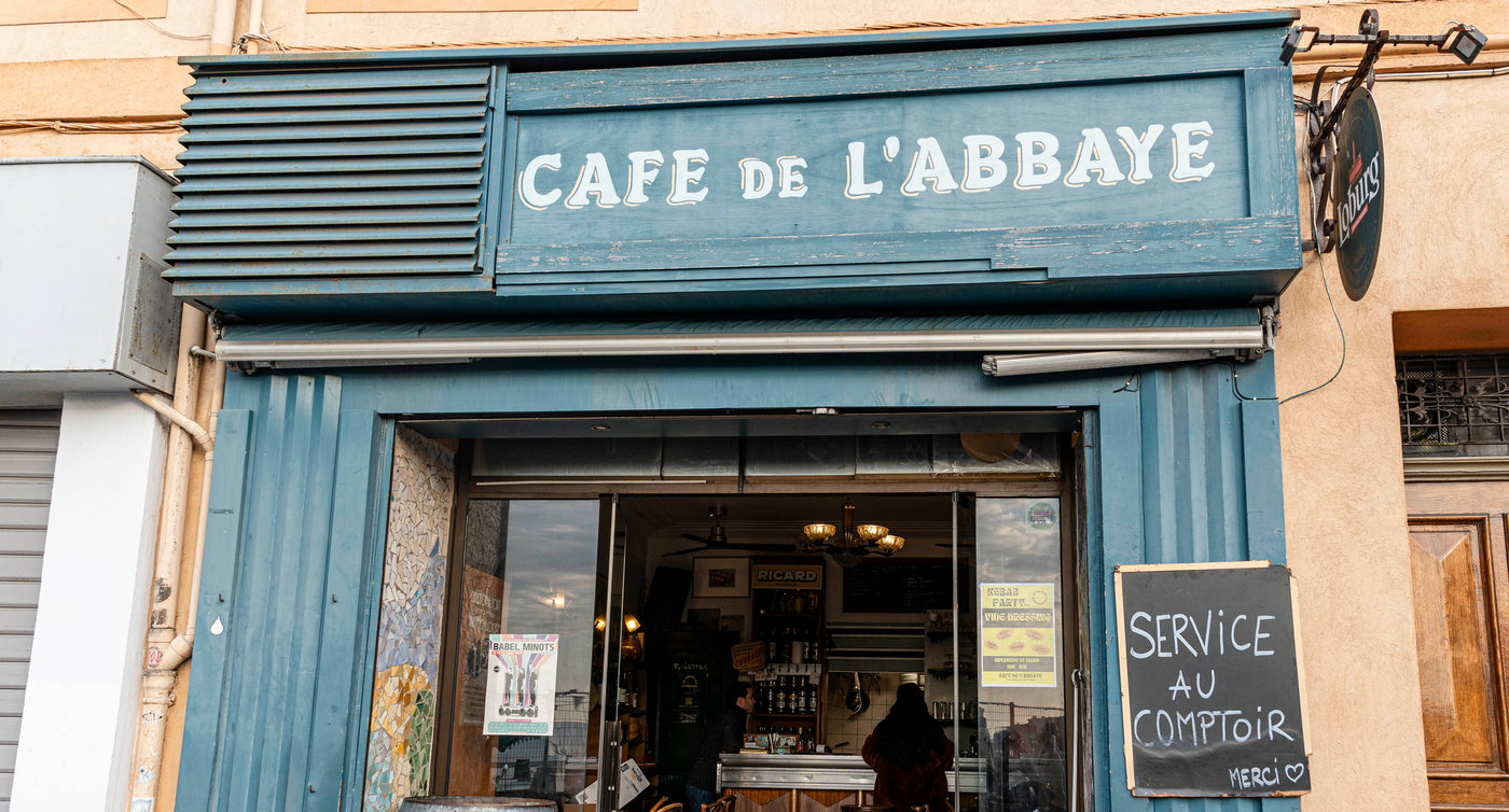 Vorderansicht des Café de l'Abbaye in Marseille, mit traditioneller blauer Markise und Schildern, die freundlich zum Service am Tresen einladen.