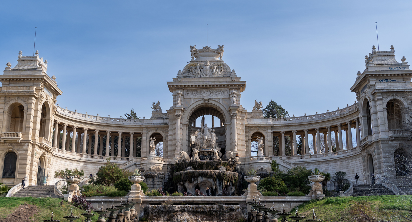 Ansicht des majestätischen Palais Longchamp in Marseille, mit seiner beeindruckenden Fontäne, umgeben von eleganten Säulengängen und Skulpturen unter klarem blauem Himmel.