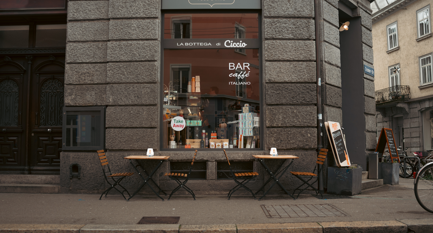 Sul marciapiede, davanti a un ristorante, ci sono tavoli e sedie di legno. Il ristorante è costruito in pietra di colore grigio scuro. 