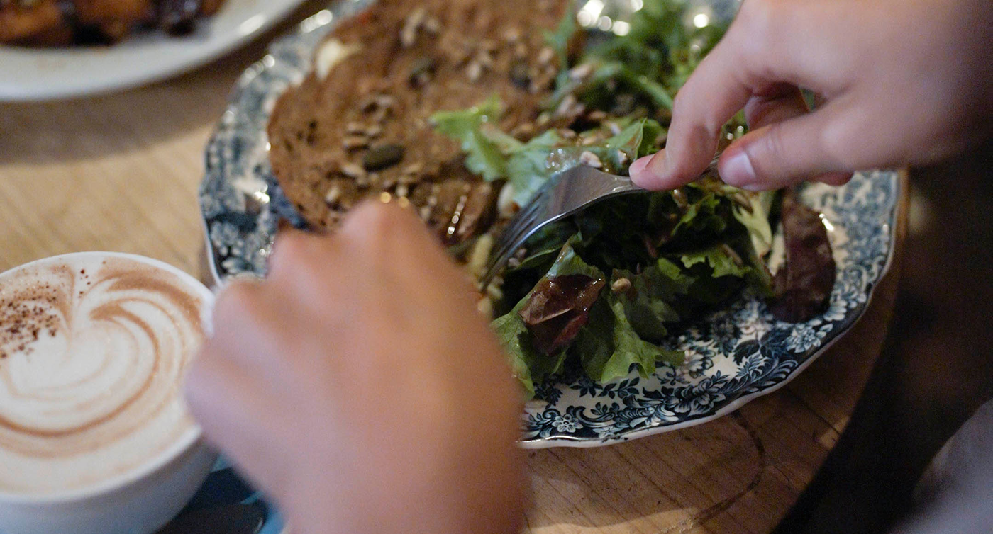 Eine Hand, die einen Salat neben einem kunstvoll angerichteten Toast mit Käse undgetrockneten Tomaten zubereitet, neben einer Tasse Cappuccino mit Herz-Schaumkrone.