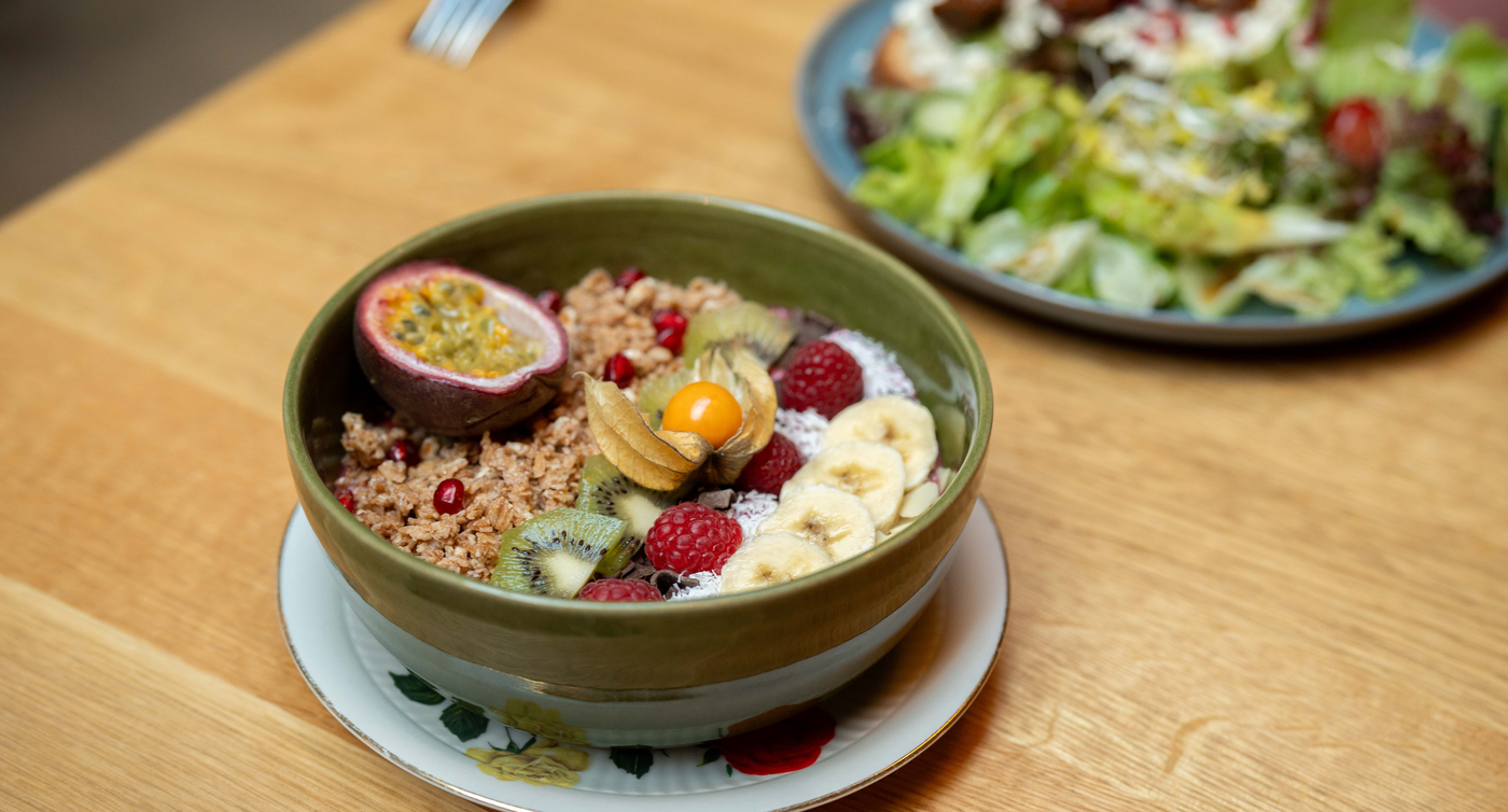 Ein grüner Teller mit Müsli, Obst und Nüssen auf einem Holztisch mit einem Salat im Hintergrund. 