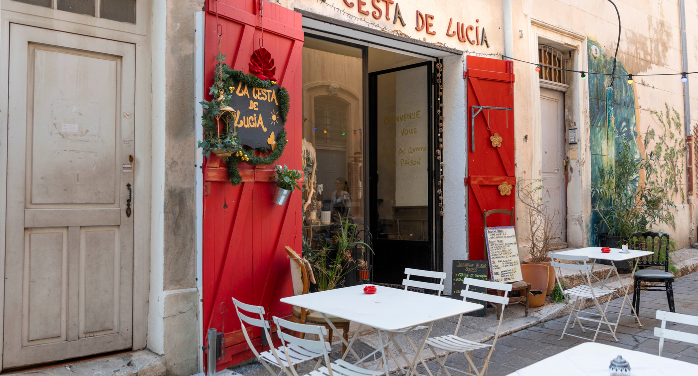 Das Restaurant 'La Cesta de Lucía' in einer ruhigen Straße von Le Panier, Marseille, mit Außensitzgelegenheiten, festlicher Türdekoration und einer Reihe bunter Lichter.
