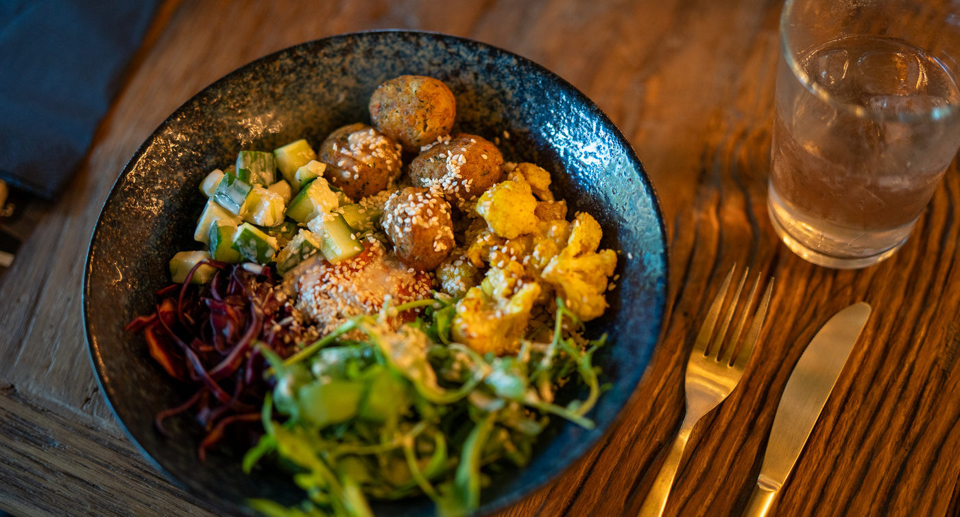 Eine Schüssel Salat auf einem Holztisch in einem Restaurant. Die Schüssel ist dunkelblau mit einer gesprenkelten Textur. Der Salat besteht aus gemischtem Grün, Rotkohl, Gurken und Falafelbällchen. Die Falafelbällchen sind goldbraun und sehen knusprig aus. Der Salat ist mit Sesamsamen und einem cremigen Dressing garniert. Neben der Schüssel stehen ein Glas Wasser und eine goldene Gabel und ein Messer. 