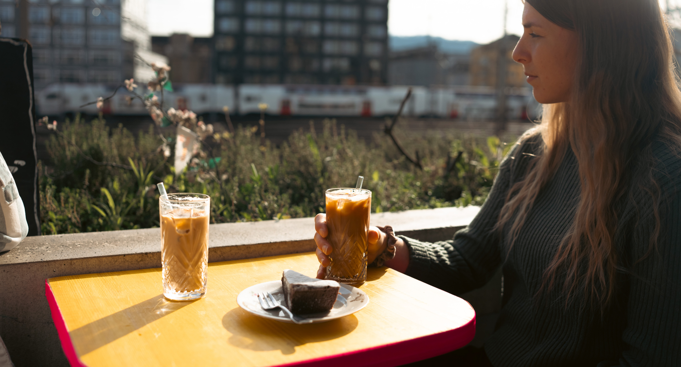 Une femme assise à une table buvant un café glacé. Elle regarde par-dessus son épaule droite en direction de la voie ferrée.  