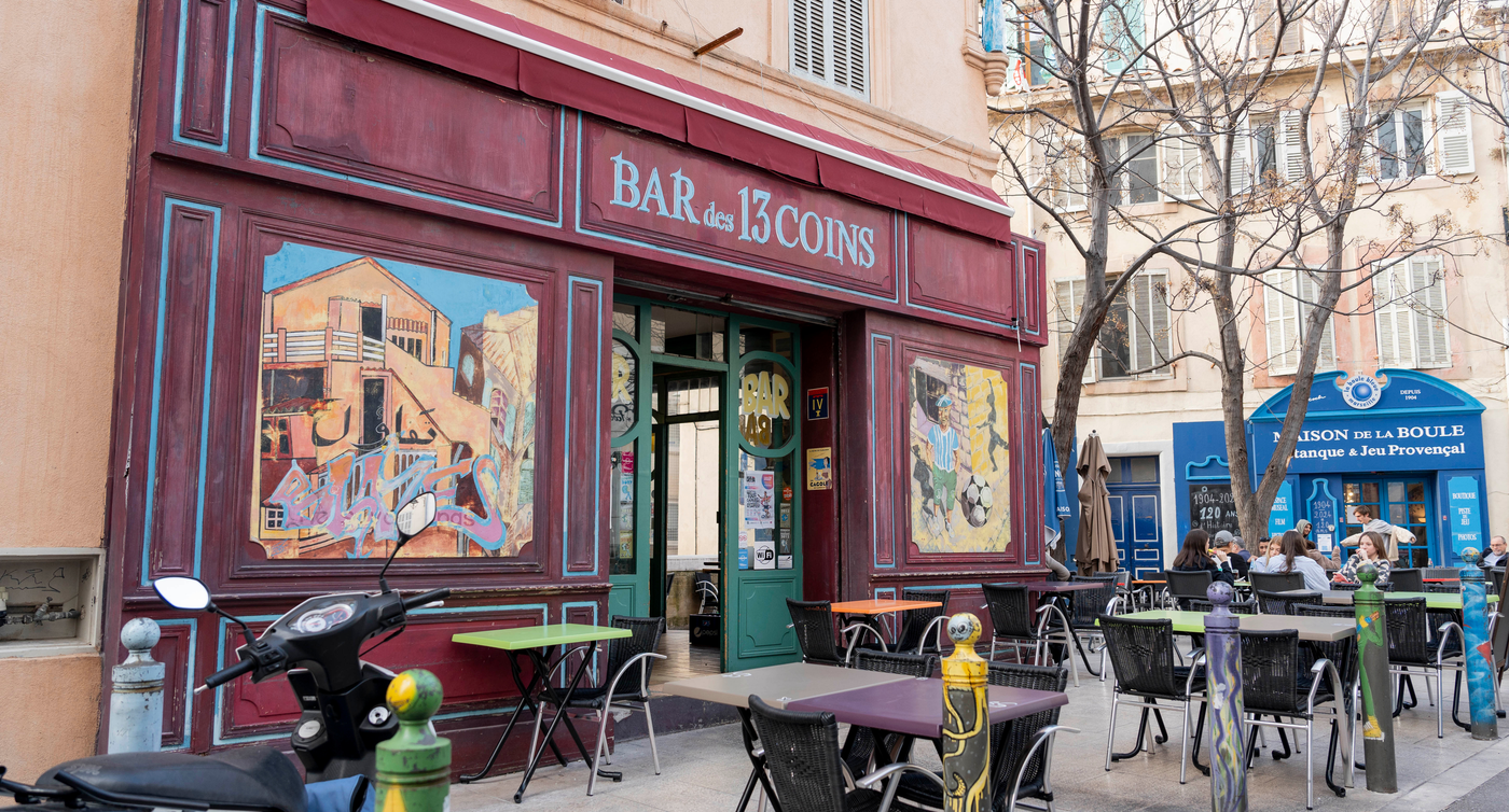Vue extérieure du Bar des 13 coins à Marseille et de ses fresques murales colorées, au milieu de terrasses et d’arbres, par un jour ensoleillé.