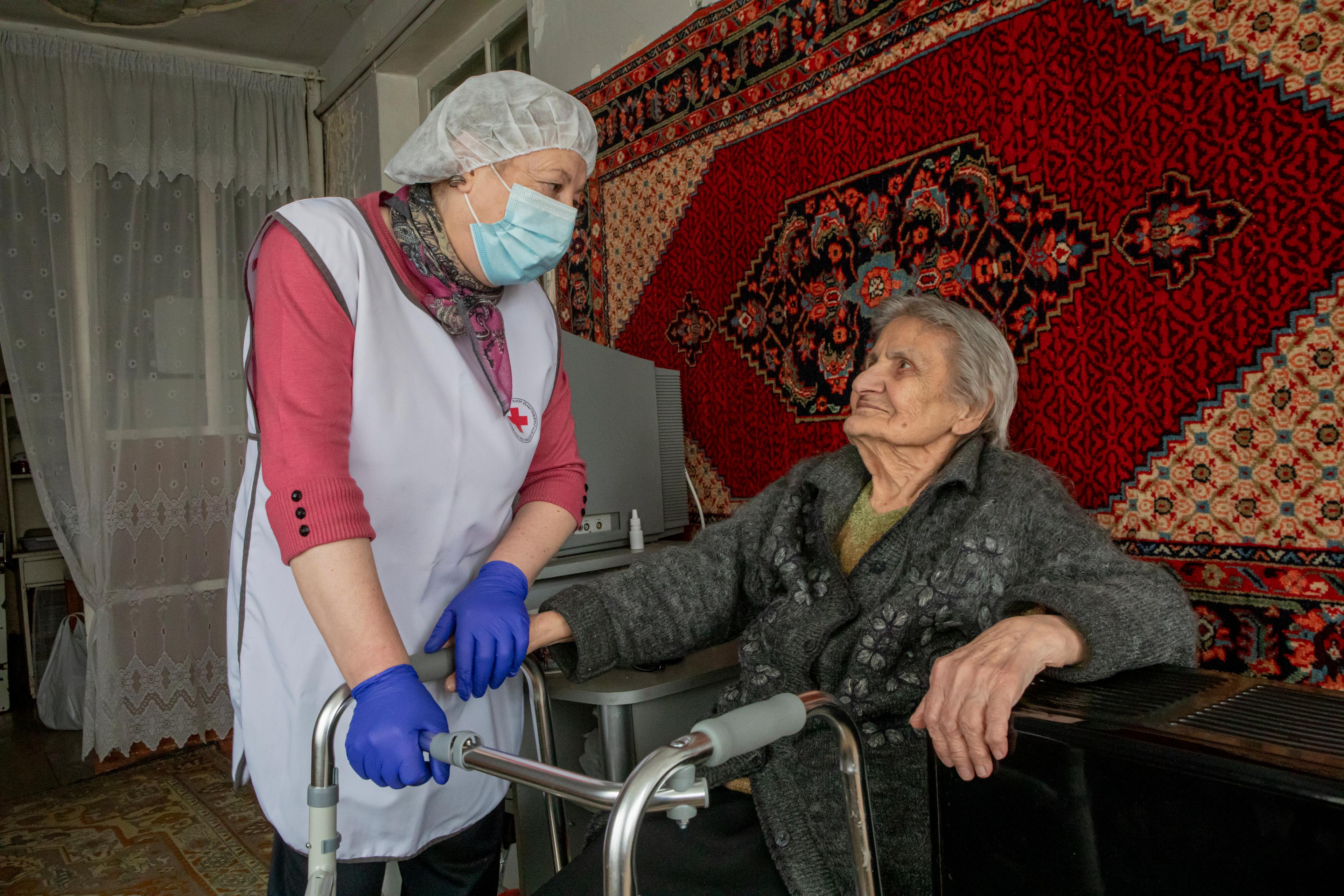 Una signora che riceve cure è seduta sul deambulatore in casa sua e guarda sorridendo l’operatrice sanitaria alla sua sinistra.