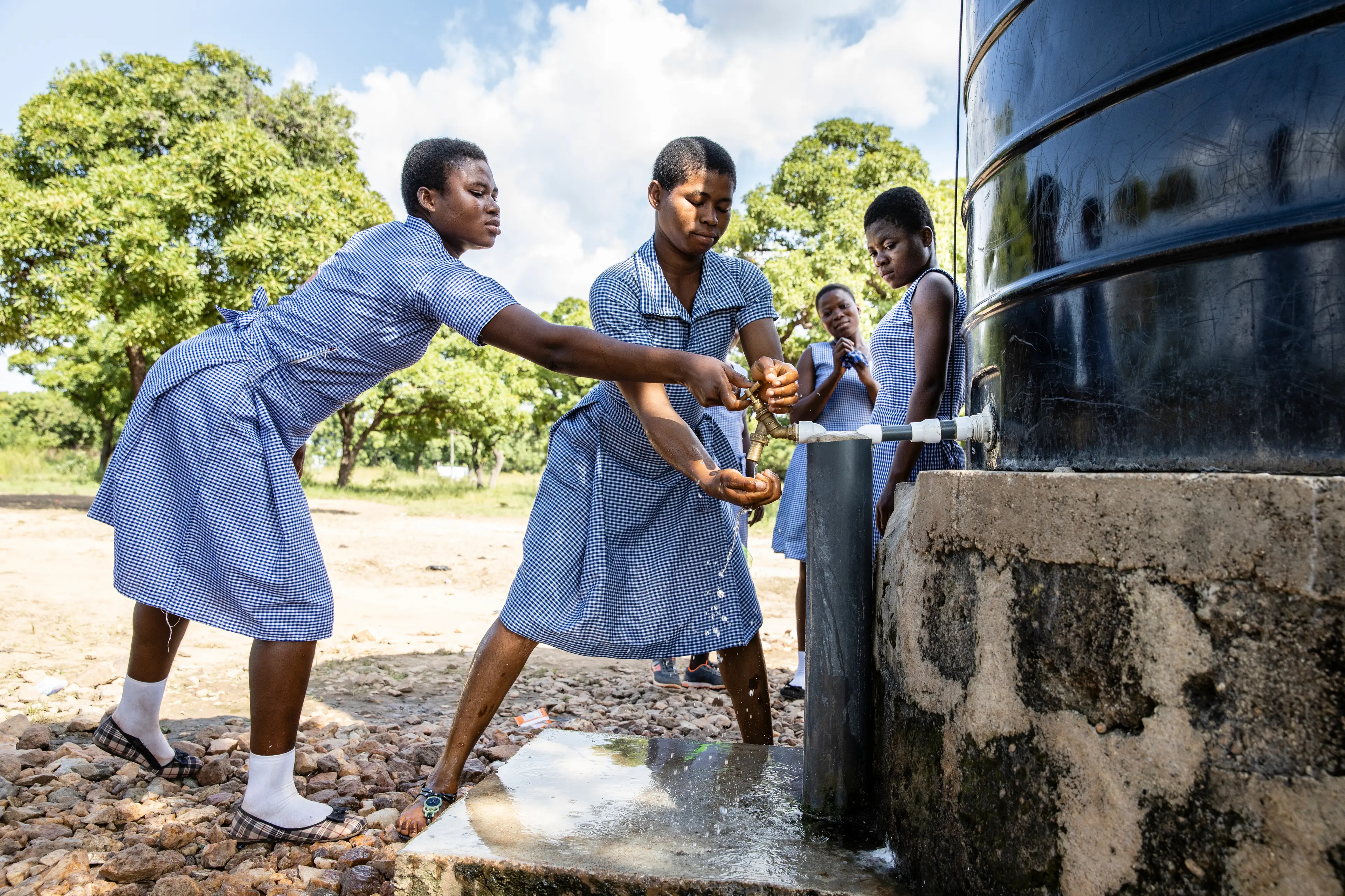 Schoolgirls washing their hands in front of a water tank. They all wear a school uniform.