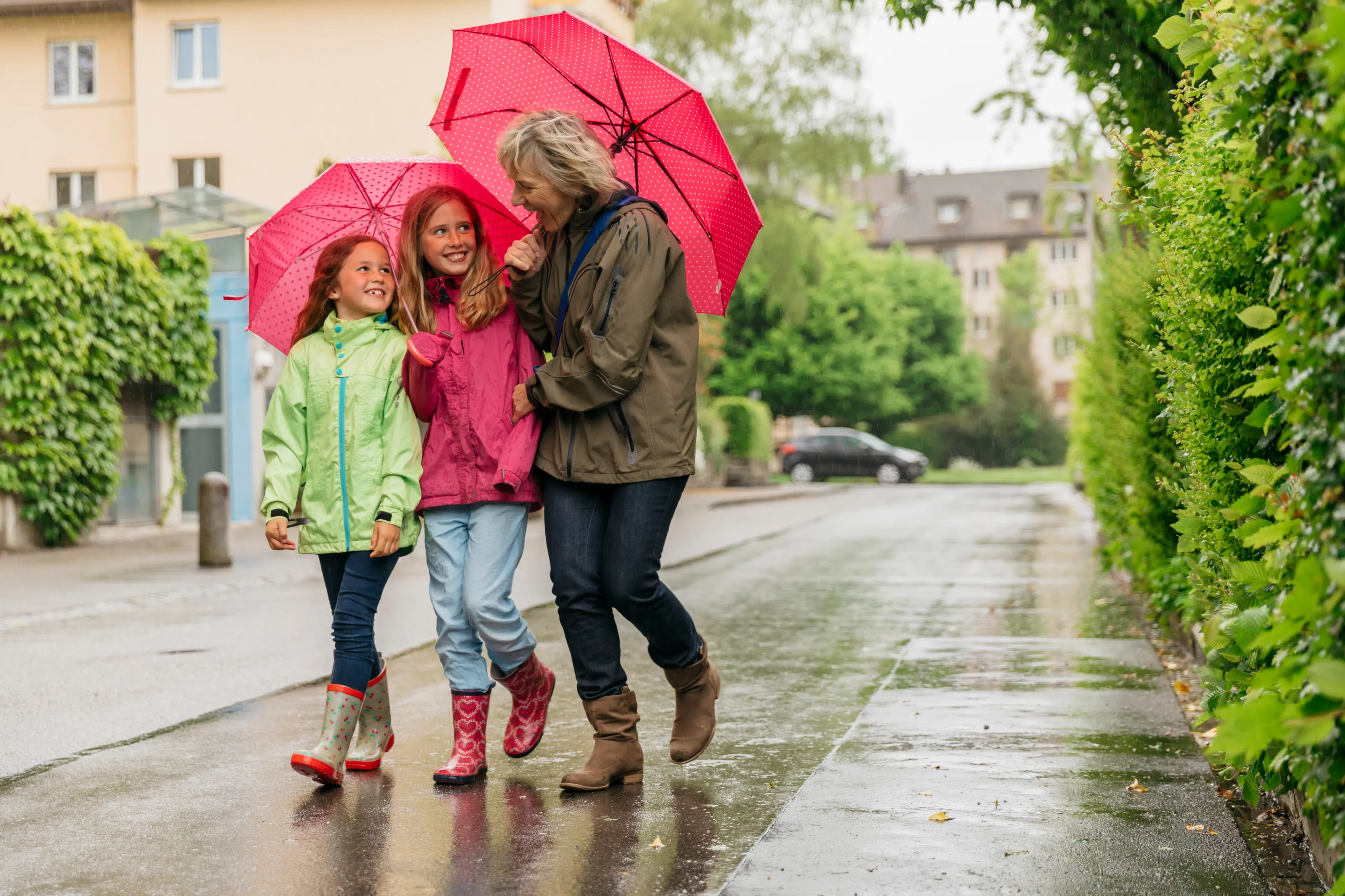 A grandmother and two little girls shelter under two pink umbrellas walking in the rain and joking.