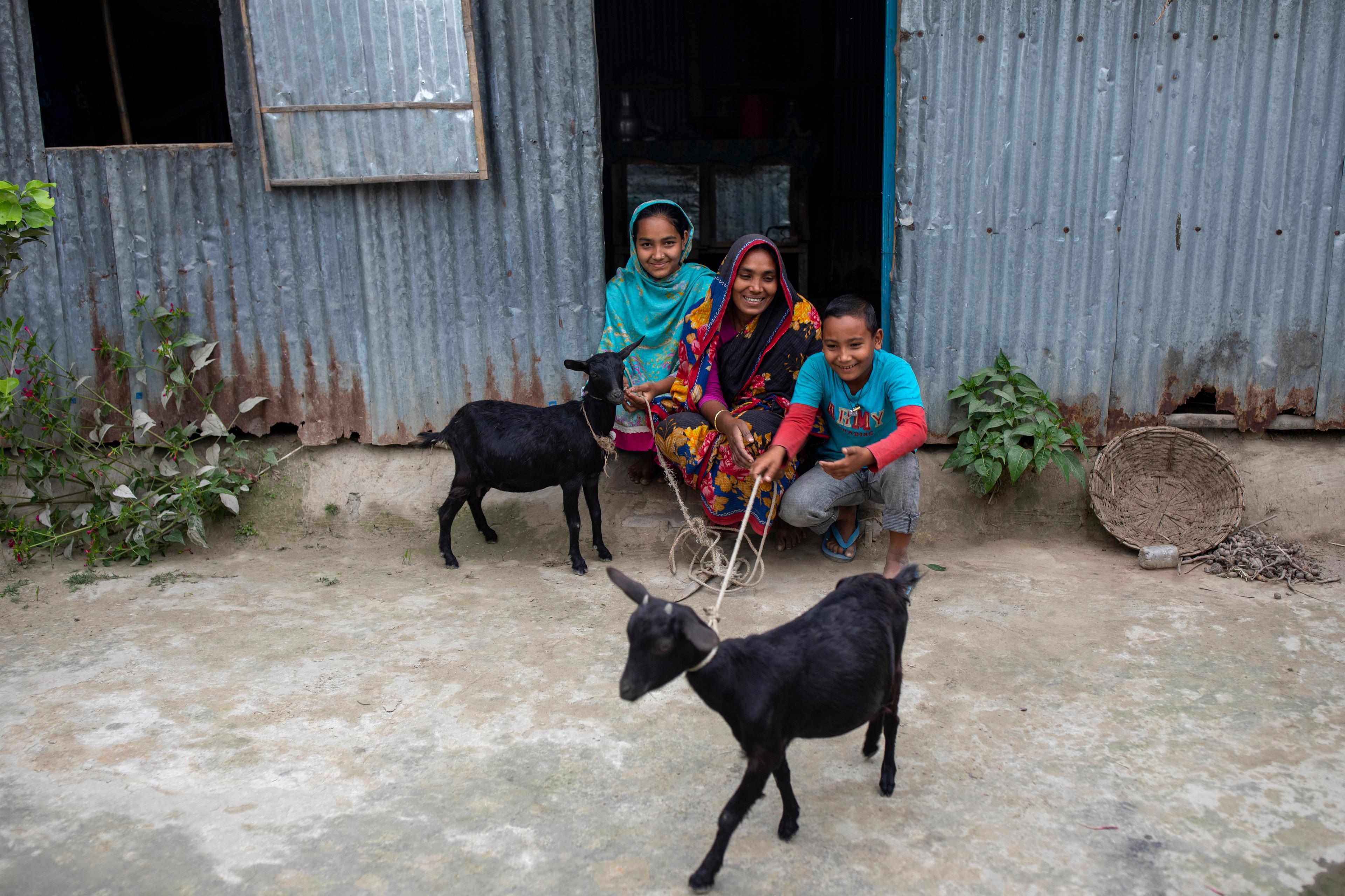 Une femme et deux enfants sont assis devant une maison en tôle ondulée et rient aux éclats. A côté d'eux se trouvent deux chèvres. 